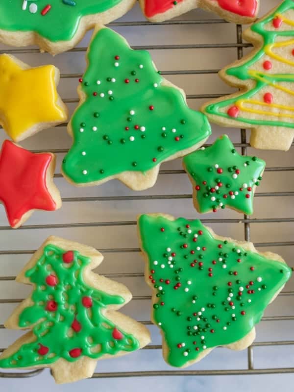 Assorted festive sugar cookies in the shapes of Christmas trees and stars, decorated with a sugar cookie icing recipe in green, red, and yellow hues, plus colorful sprinkles, resting on a cooling rack.