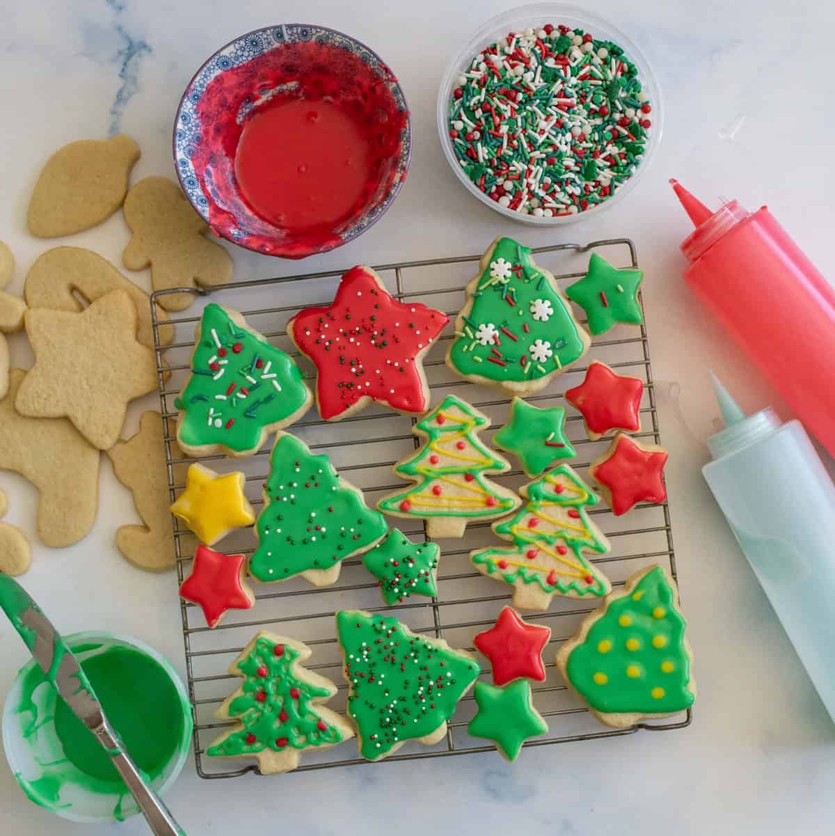 A variety of Christmas cookies shaped like trees, stars, and stockings are decorated with colorful red, green, and white icing and sprinkles. They are placed on a cooling rack, with bowls of icing and more sprinkles nearby.