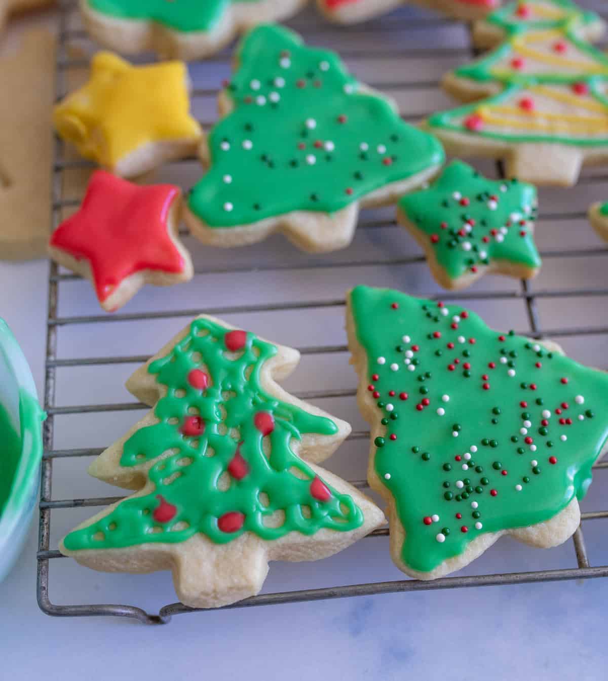 A cooling rack holds Christmas-themed cookies shaped like trees and stars. The cookies are decorated with bright green icing and multicolored sprinkles. Some are red and yellow stars, adding a festive touch.