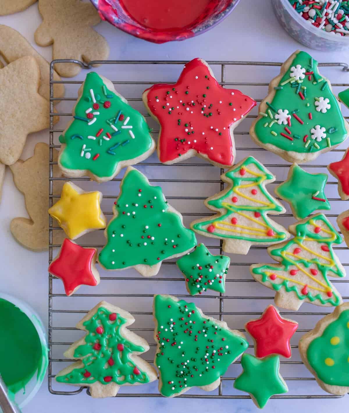 A cooling rack with colorful Christmas cookies shaped like trees and stars, decorated with red, green, and yellow icing and sprinkles. Nearby icing bowls are seen, adding to the festive feel of the scene.