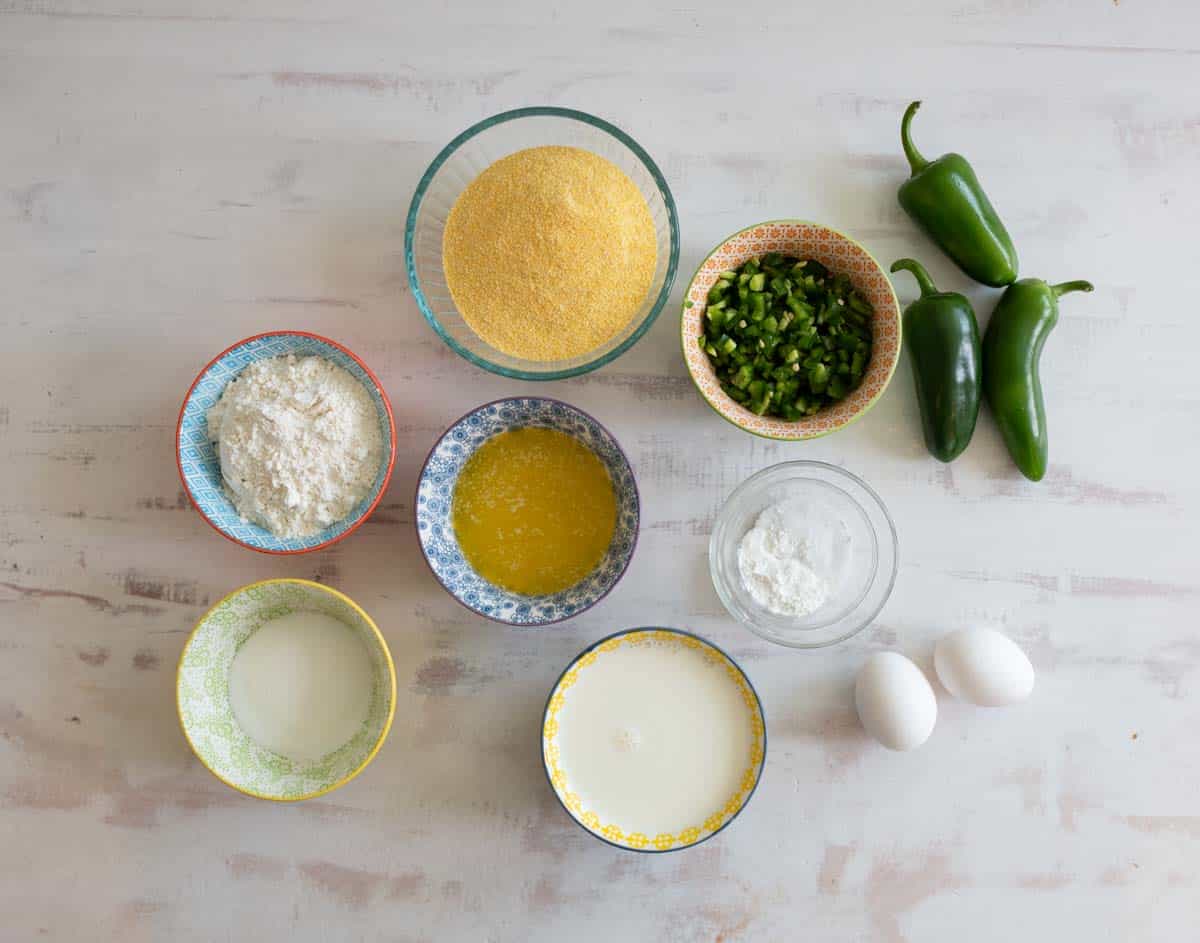 Various ingredients for cornbread are neatly arranged on a white surface, including cornmeal, chopped green peppers, jalapeños, flour, melted butter, baking powder, milk, and two eggs in colorful bowls.