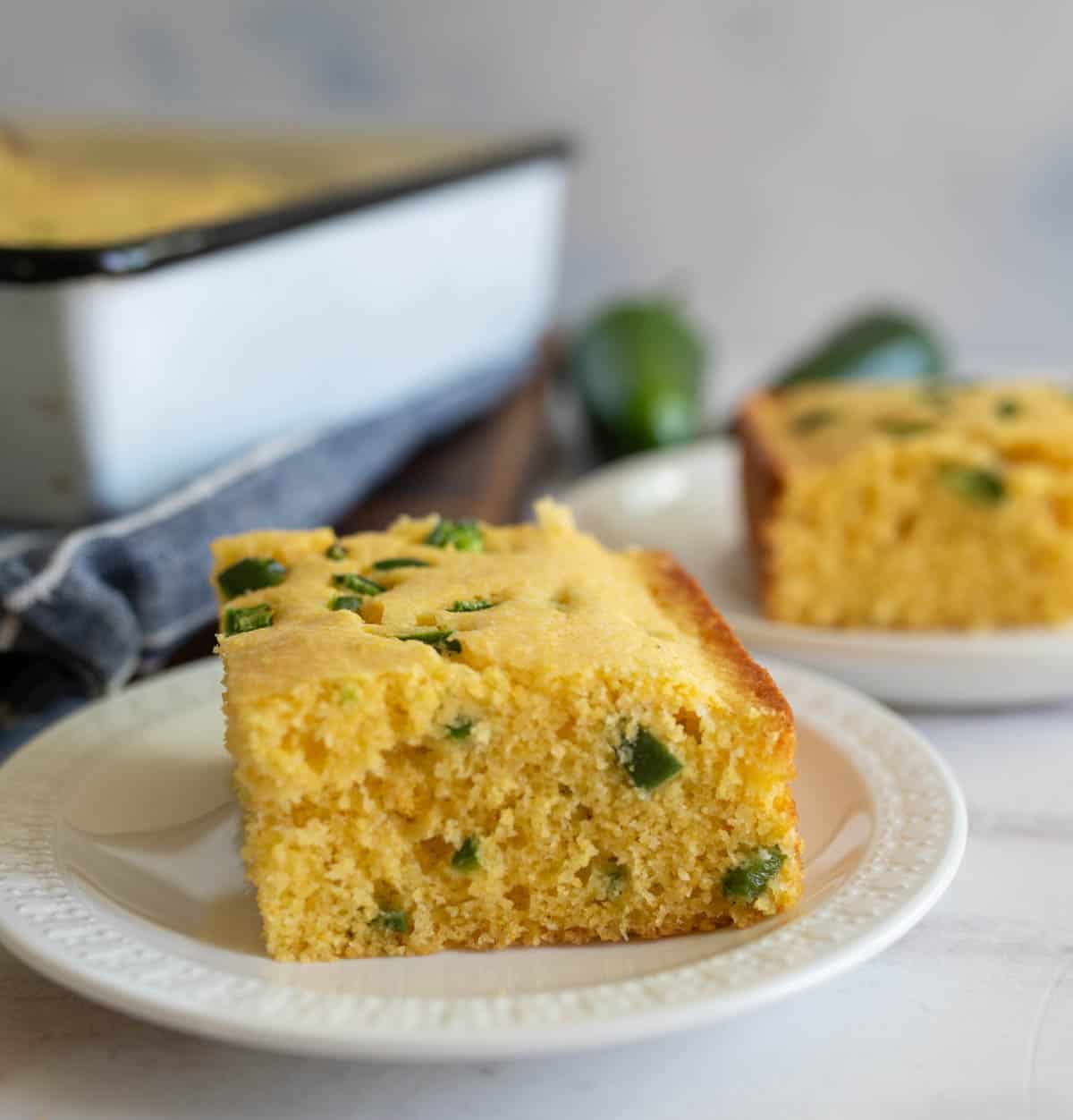 A slice of cornbread with jalapeño pieces on a white plate. Another slice is in the background, with a baking tray partially visible. The cornbread is golden and moist, set on a light, neutral surface.