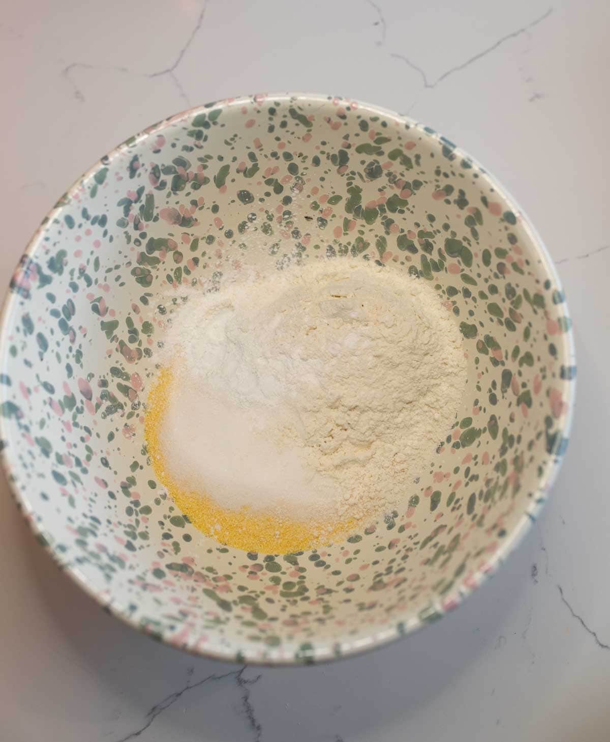 A speckled mixing bowl contains a mound of dry ingredients, including flour and sugar, on a light-colored countertop.