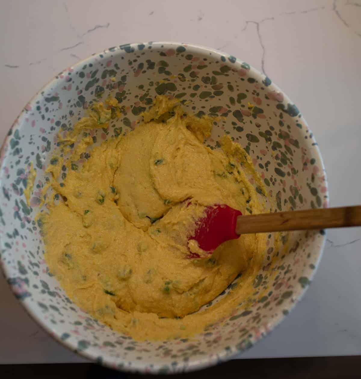 A colorful speckled bowl on a marble countertop contains thick, yellow batter mixed with green ingredients. A red spatula with a wooden handle is resting in the batter.