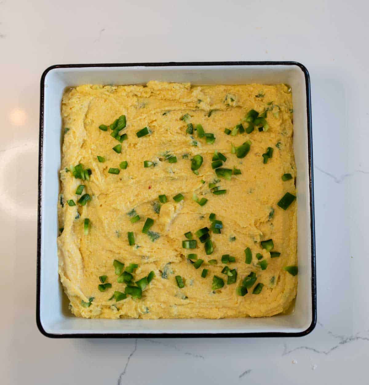 Baking pan with uncooked cornbread batter topped with chopped green jalapeños, placed on a white marble countertop.