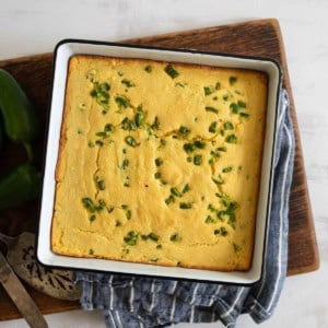 A square pan of freshly baked jalapeno cornbread topped with chopped green peppers sits on a wooden board. A blue-striped cloth and a serving utensil are nearby.