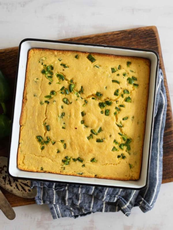 A square pan of freshly baked jalapeno cornbread topped with chopped green peppers sits on a wooden board. A blue-striped cloth and a serving utensil are nearby.