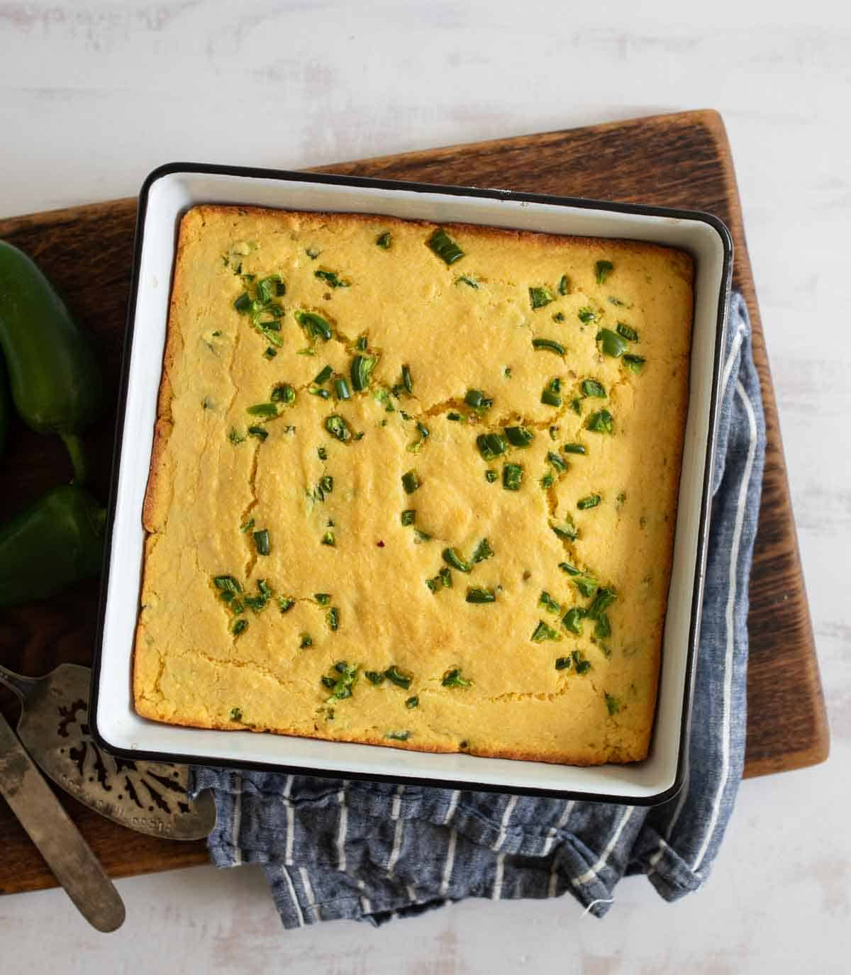 A square pan of freshly baked cornbread topped with chopped green jalapeños sits on a wooden board. A striped blue cloth is underneath, and a metal serving utensil is nearby.