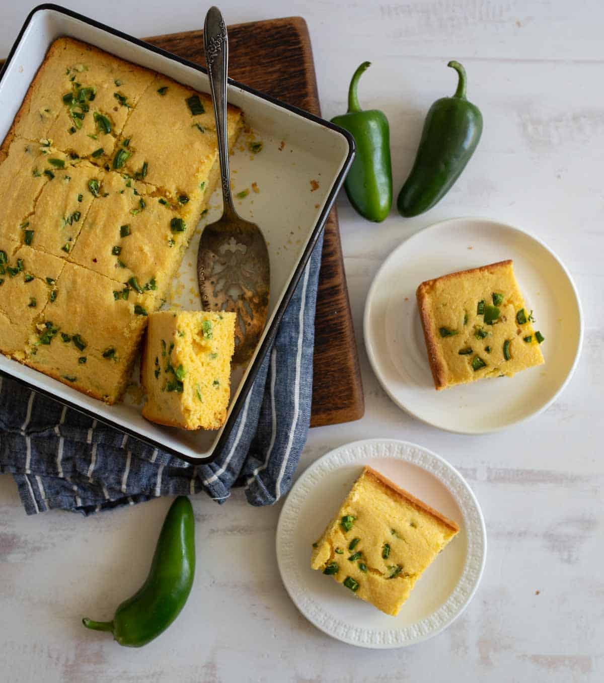 A baking dish with sliced jalapeño cornbread on a wooden board. Two pieces of cornbread are on separate white plates nearby. Whole jalapeños and a blue-striped cloth are also in the scene.