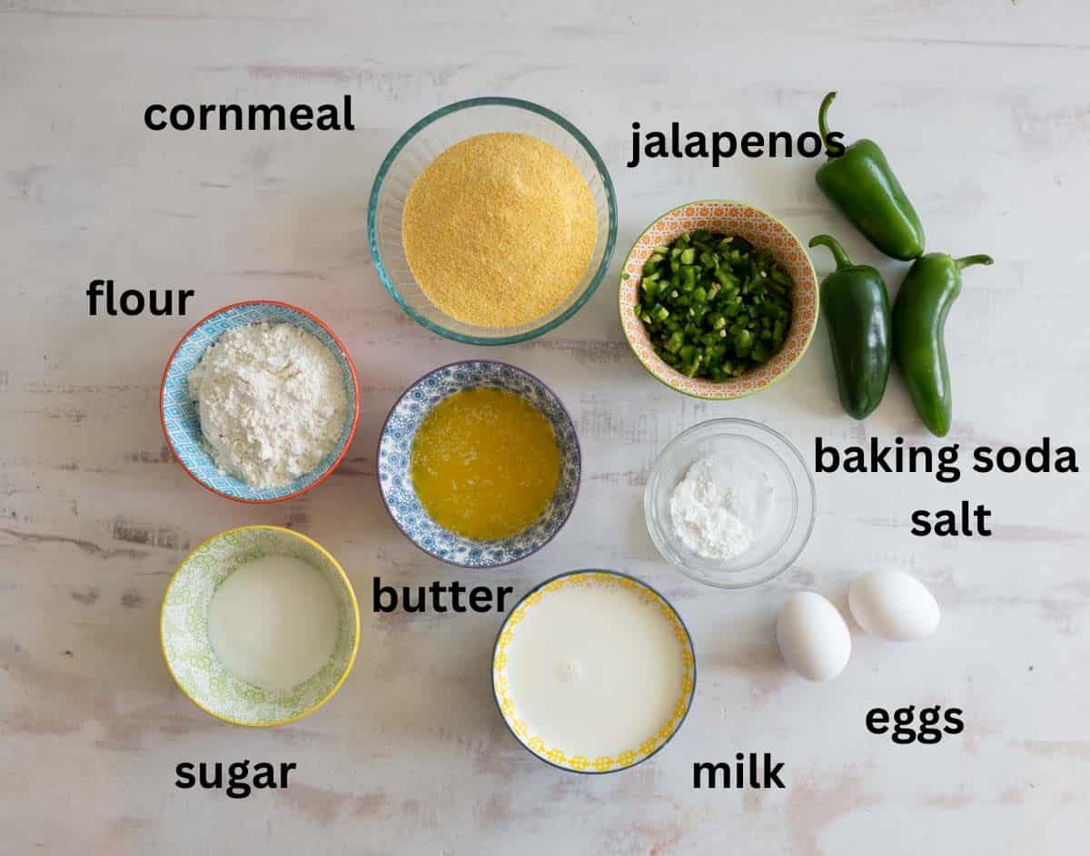 Ingredients for cornbread are arranged on a table: cornmeal, jalapenos, flour, baking soda, salt, sugar, butter, milk, and eggs. Each ingredient is in a bowl or cup and labeled accordingly.