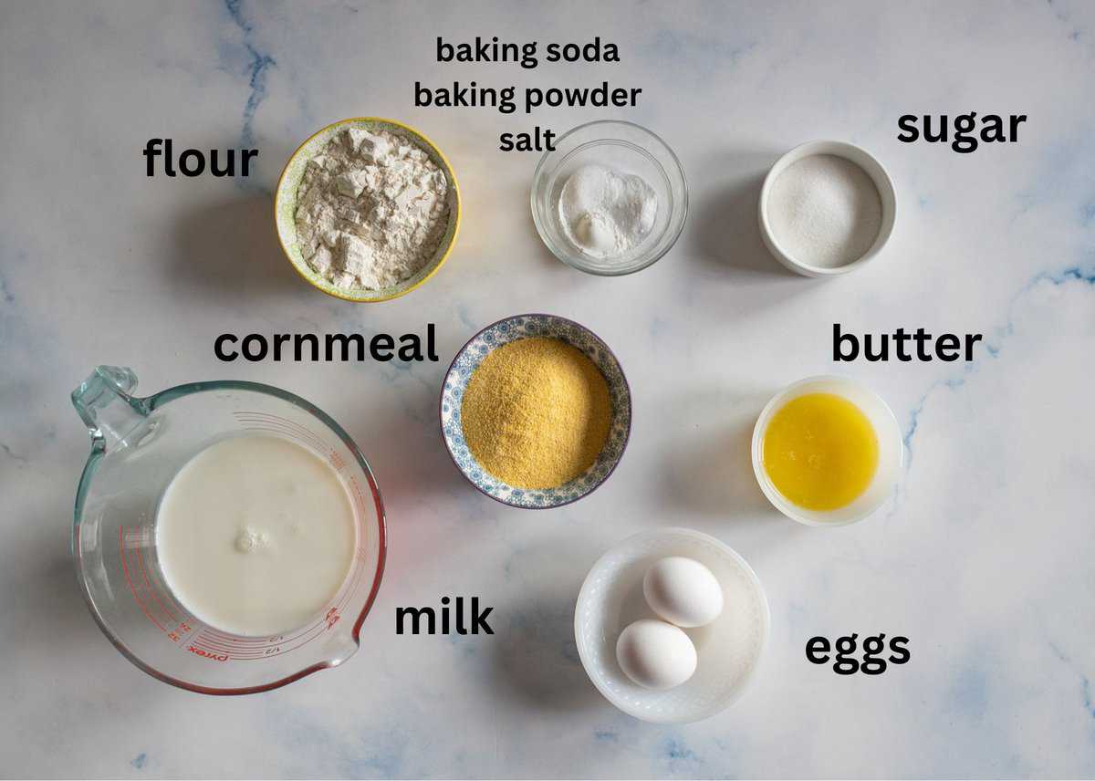 Top view of ingredients labeled for cornbread: flour, sugar, baking soda, baking powder, salt, cornmeal, melted butter, milk in a measuring cup, and two eggs, all arranged on a marble surface.