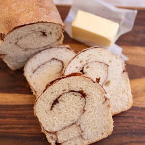 baked loaf of cinnamon bread with 3 slices and butter in the background on brown board.