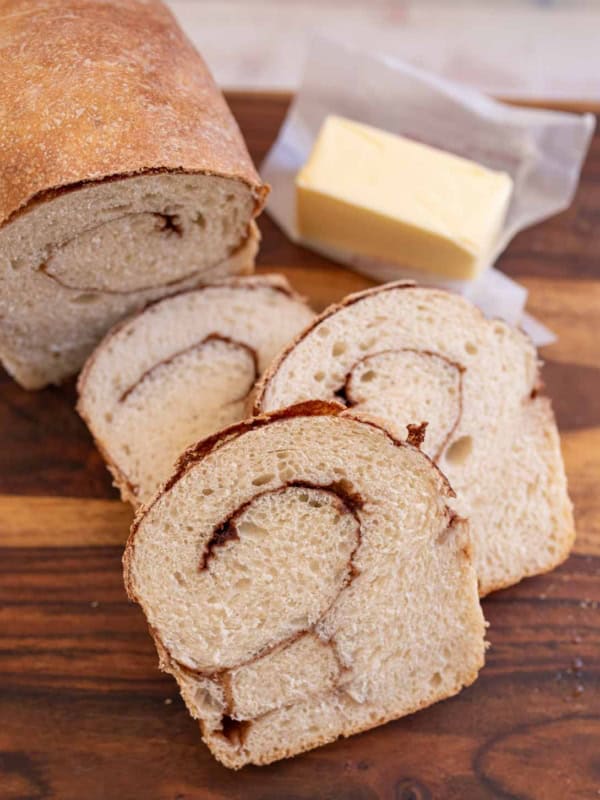 baked loaf of cinnamon bread with 3 slices and butter in the background on brown board.