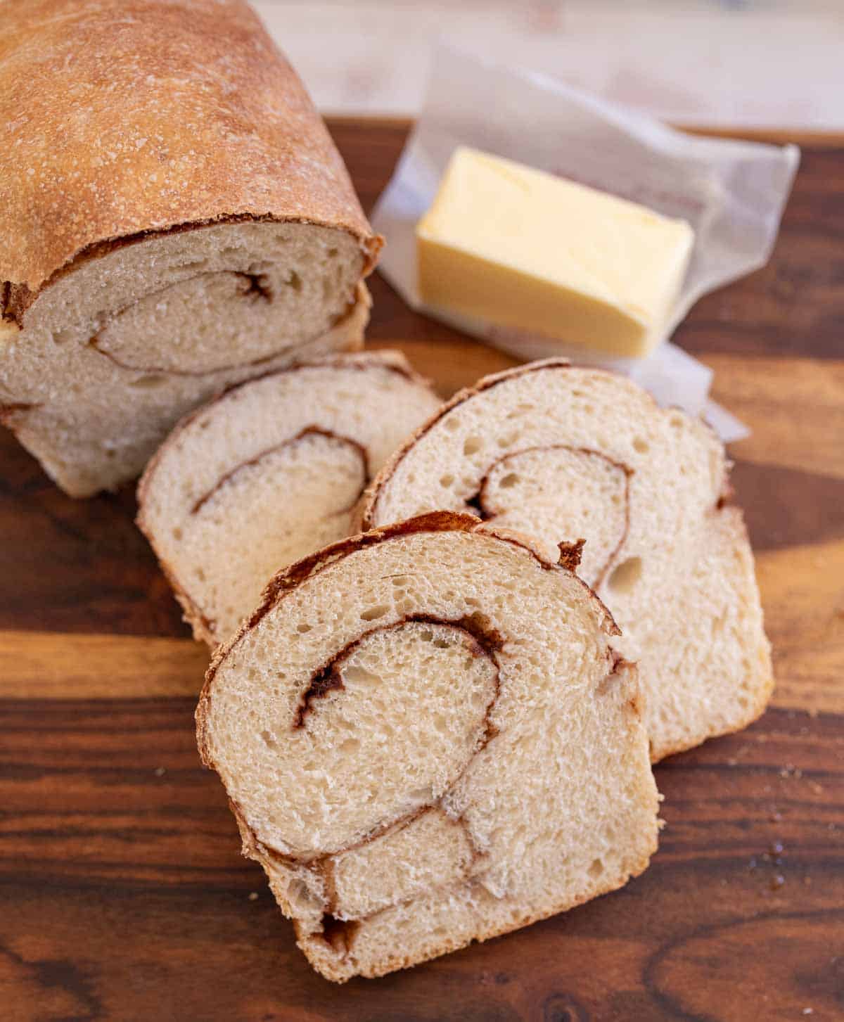 baked loaf of cinnamon bread with 3 slices and butter in the background on brown board.