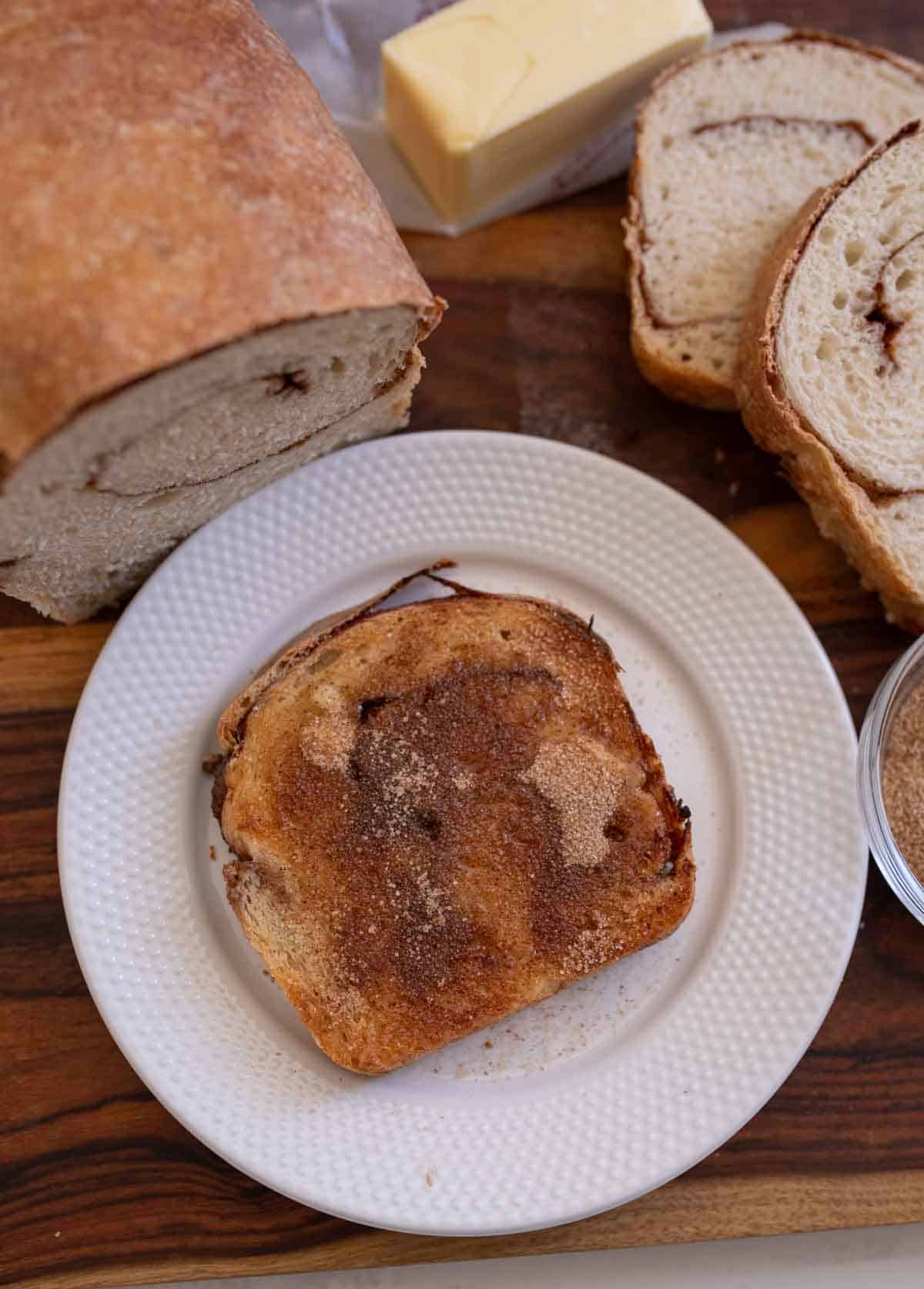 slice of cinnamon swirl bread on white plate with other slices and rest of loaf and butter in background.