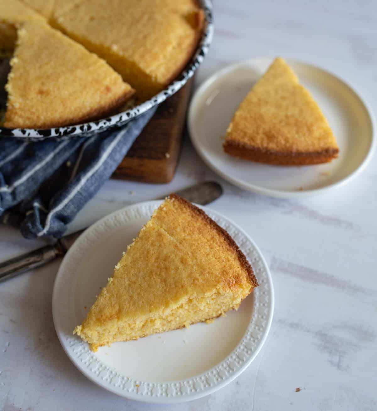 Two slices of cornbread are served on white plates next to a larger pan with a partially cut cornbread. A blue cloth is folded nearby, and a knife rests on the table, all on a light marble surface.