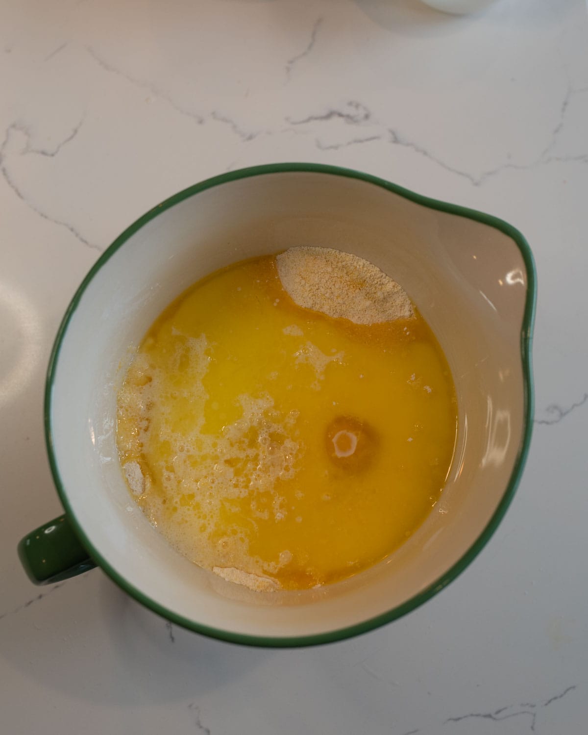 A green ceramic mixing bowl containing melted butter and dry ingredients on a marble countertop, ready to be mixed for baking.