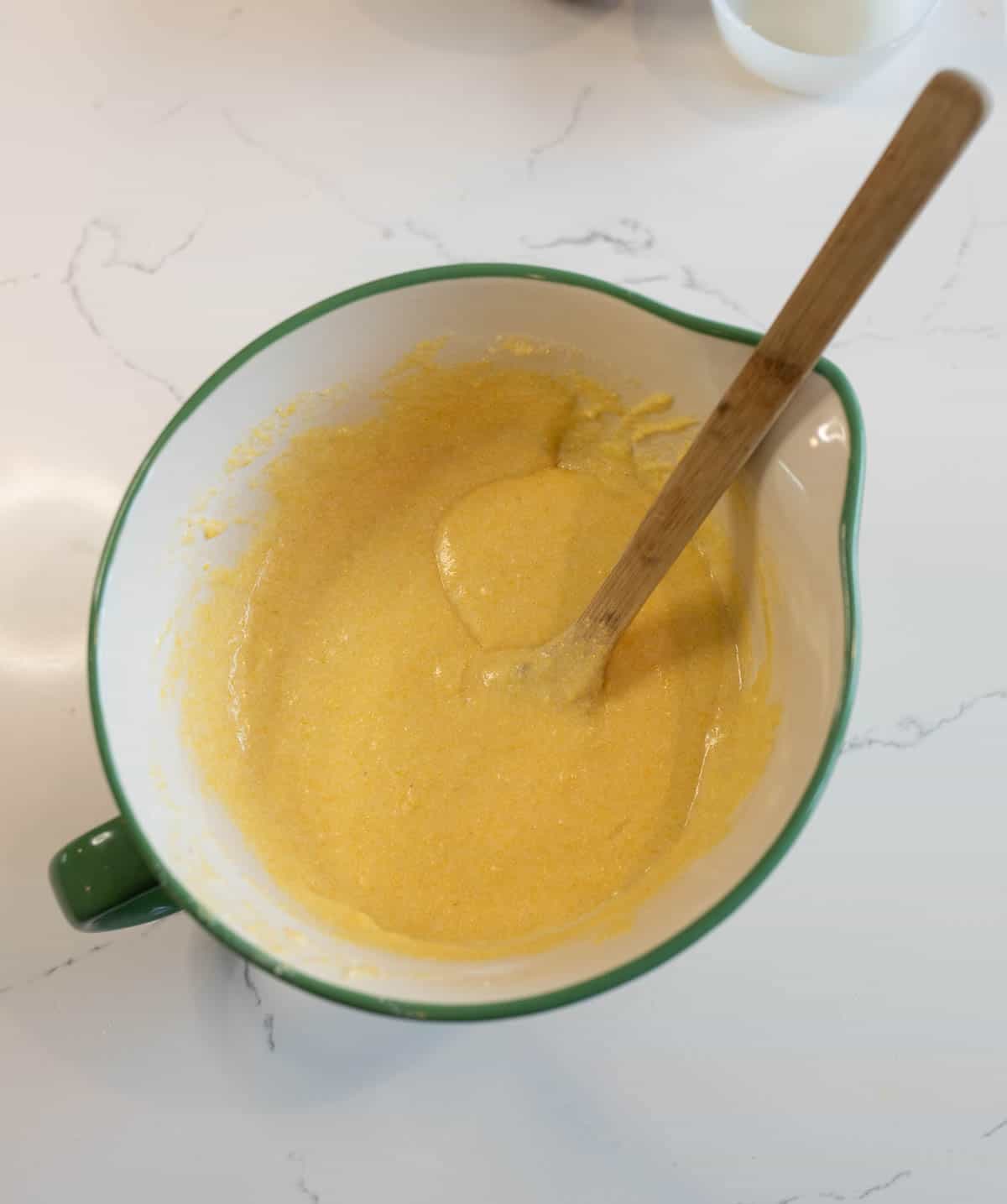 A green mixing bowl filled with a smooth, yellow batter is on a white marble countertop. A wooden spoon rests inside the bowl, partially covered by the batter.