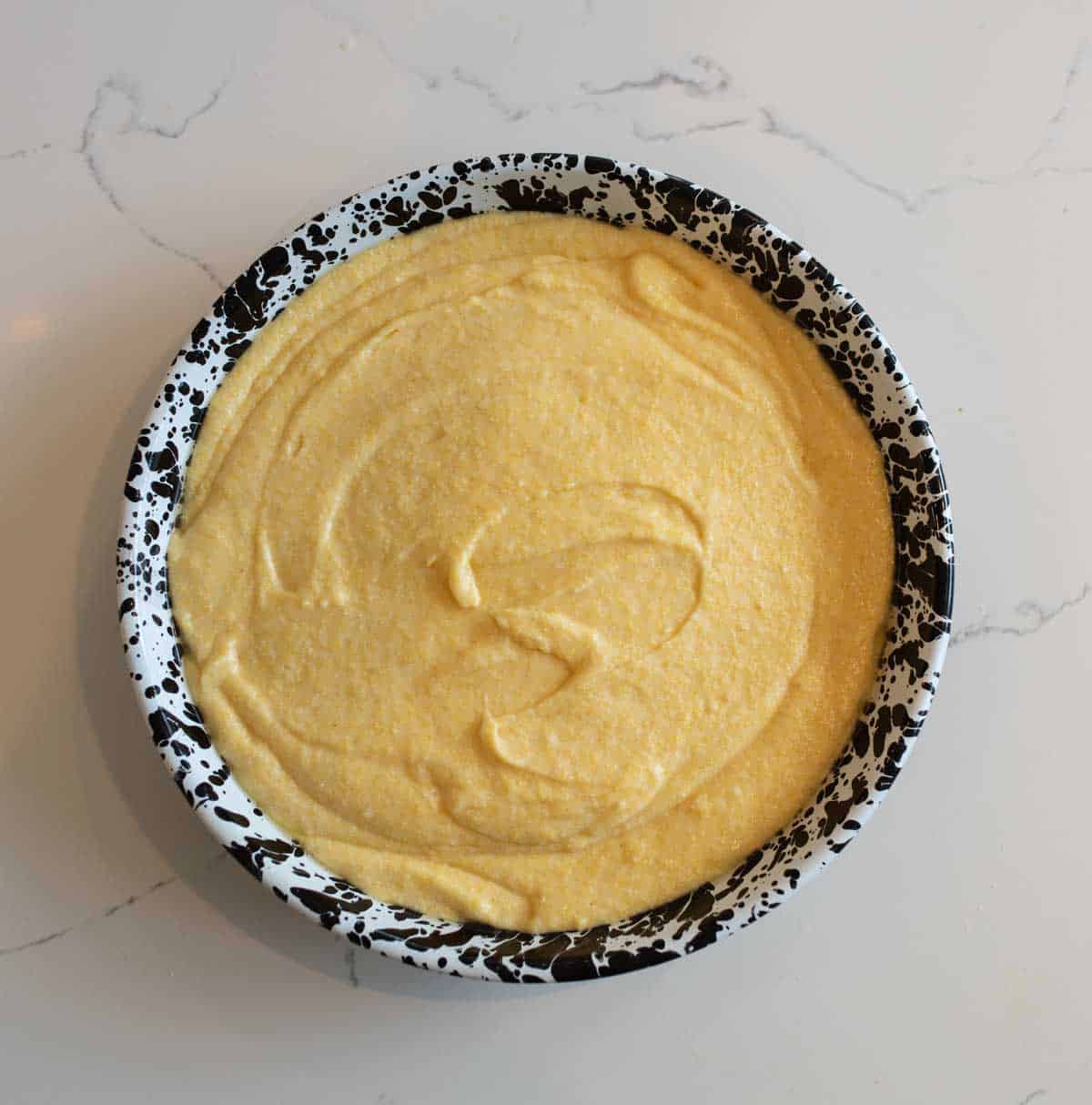 A round, black-and-white marbled baking dish filled with smooth, uncooked cornbread batter sits on a white marble countertop. The surface of the batter is swirled and ready for baking.