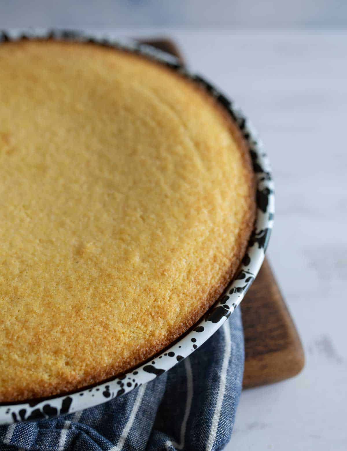 A freshly baked cornbread in a round, speckled pan rests on a blue and white striped cloth on a wooden cutting board. The cornbread has a golden-brown crust and smooth surface, indicating it is ready to be served.