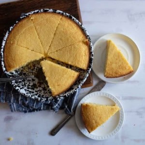 A round cornbread is partially sliced into wedges on a speckled dish, with two slices placed on separate white plates. A metal serving utensil rests in the dish on a wooden board. A folded cloth lies beneath the dish.