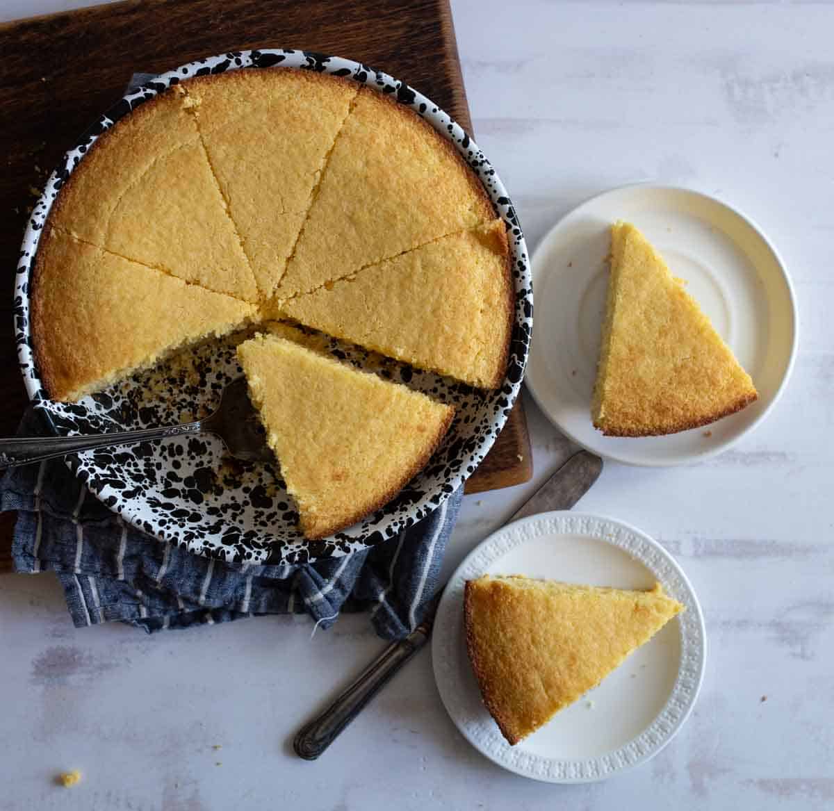 A round cornbread is partially sliced into wedges on a speckled dish, with two slices placed on separate white plates. A metal serving utensil rests in the dish on a wooden board. A folded cloth lies beneath the dish.