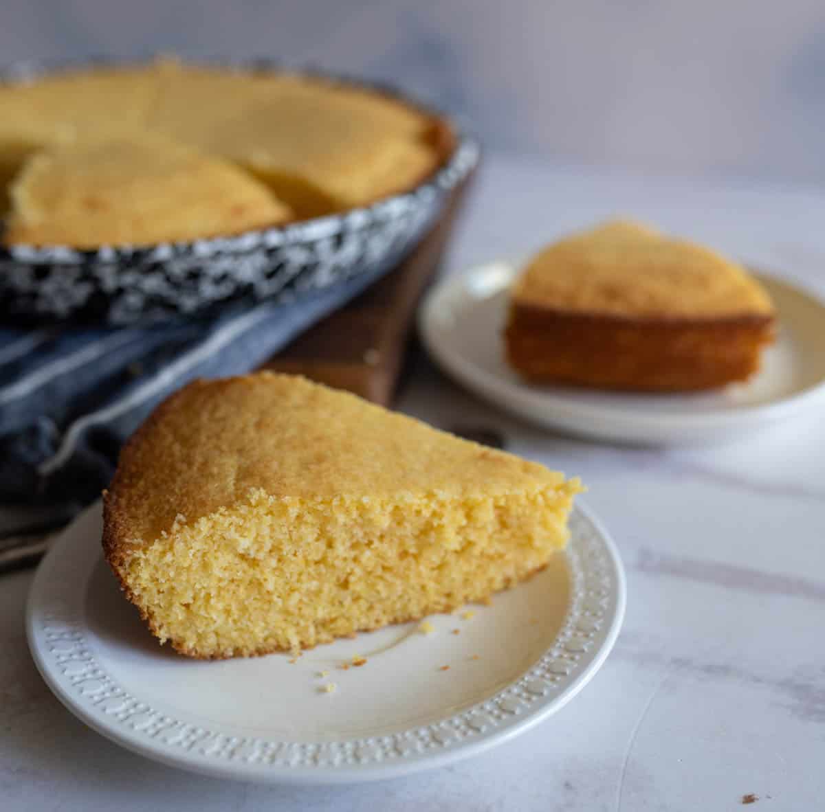 A slice of golden-brown cornbread is served on a white plate. A full cornbread loaf is visible in the background, cut into several pieces in a dark baking dish. The scene is set on a light-colored table.