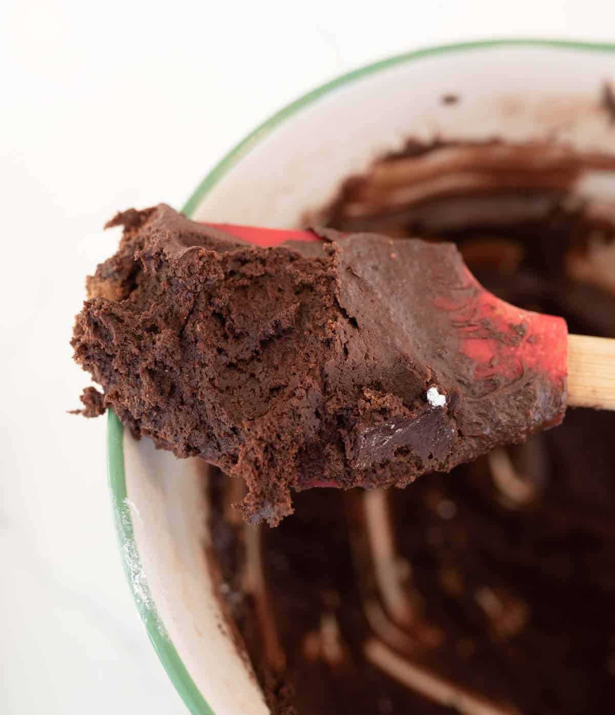 A red spatula scooping thick chocolate brownie batter from a mixing bowl. The bowl has a green rim and is partially covered in batter. The rich, dark chocolate mixture looks smooth and creamy.