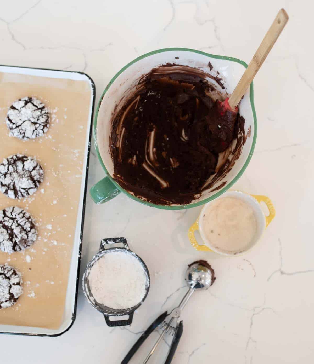 A table with a mixing bowl of chocolate dough and a spatula, a tray of baked crinkle cookies, a cup of sugar, a small bowl of flour, and an ice cream scoop on a white surface.