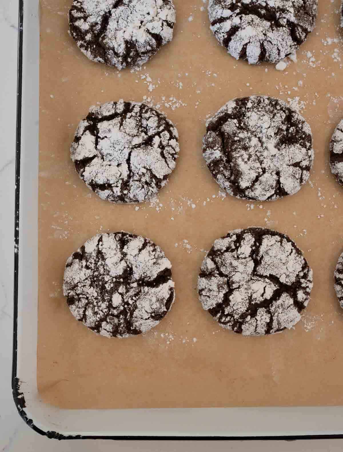 A baking sheet lined with parchment paper holds nine chocolate crinkle cookies. Each cookie is dusted with powdered sugar and has a cracked surface, creating a contrasting pattern of dark brown and white.