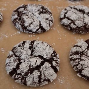 Chocolate crinkle cookies dusted with powdered sugar are displayed on a parchment-lined baking sheet. The cookies have a cracked surface, revealing the chocolate underneath.