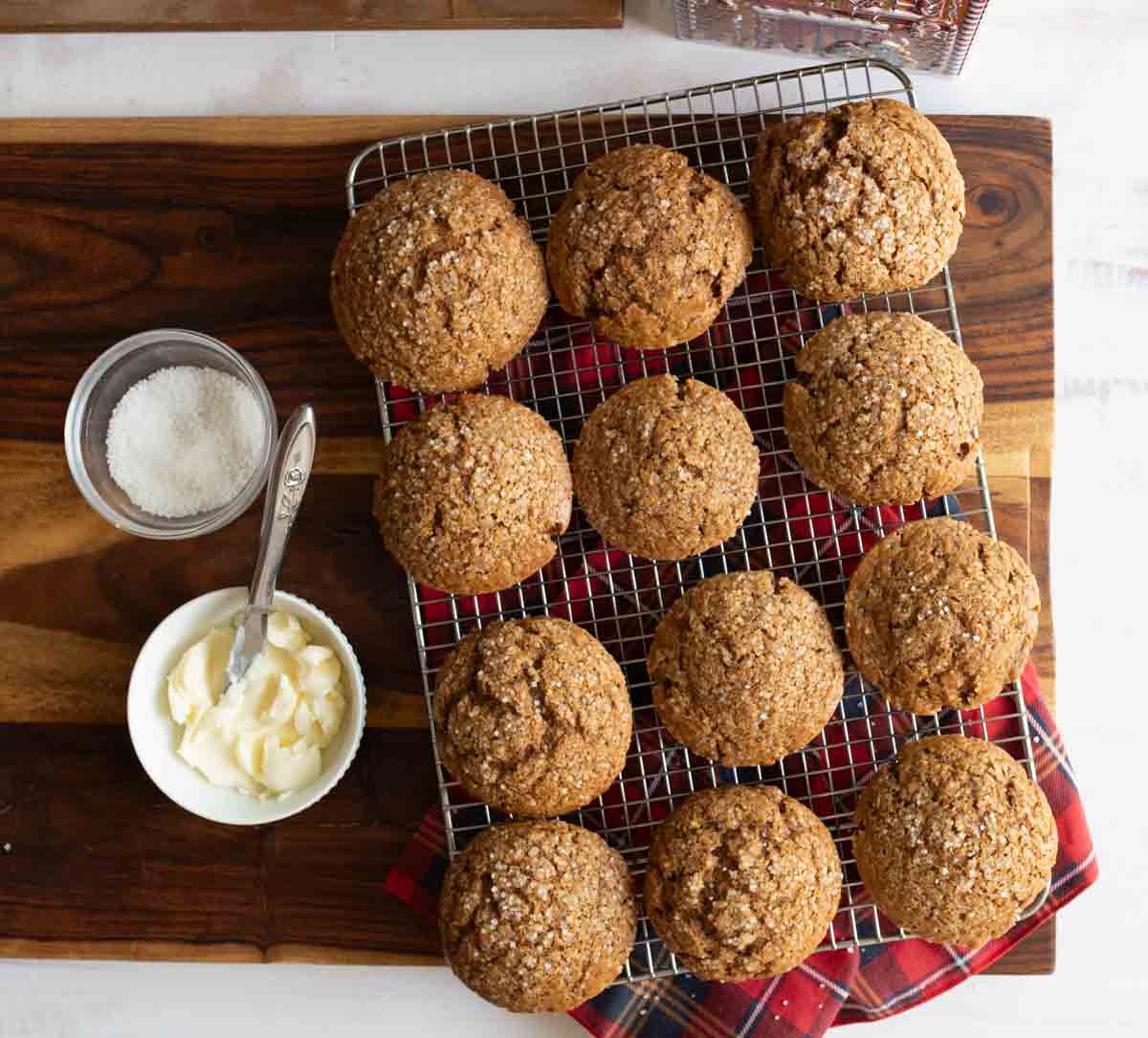 A batch of ten freshly baked muffins on a cooling rack, placed on a wooden surface. Beside the rack is a small bowl with butter and a spoon, and another bowl with sugar. A red plaid cloth is partially visible under the muffins.