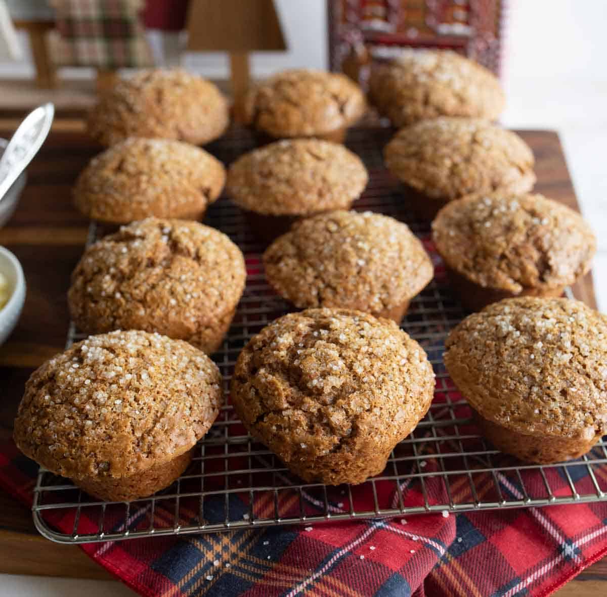 Twelve freshly baked muffins topped with sugar crystals rest on a cooling rack. Theyre placed on a red and blue plaid cloth with a wooden background, creating a cozy, homemade atmosphere.