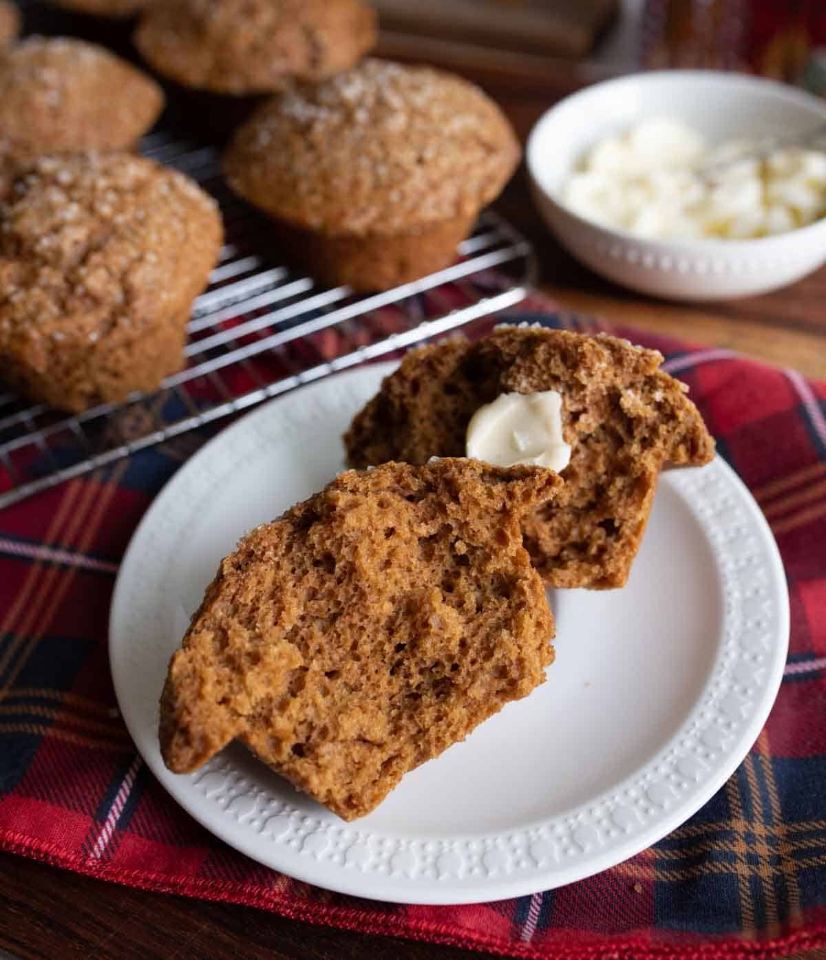 A split muffin with a dollop of butter sits on a white plate, placed on a red plaid cloth. Several whole muffins rest on a cooling rack in the background, next to a bowl of butter.