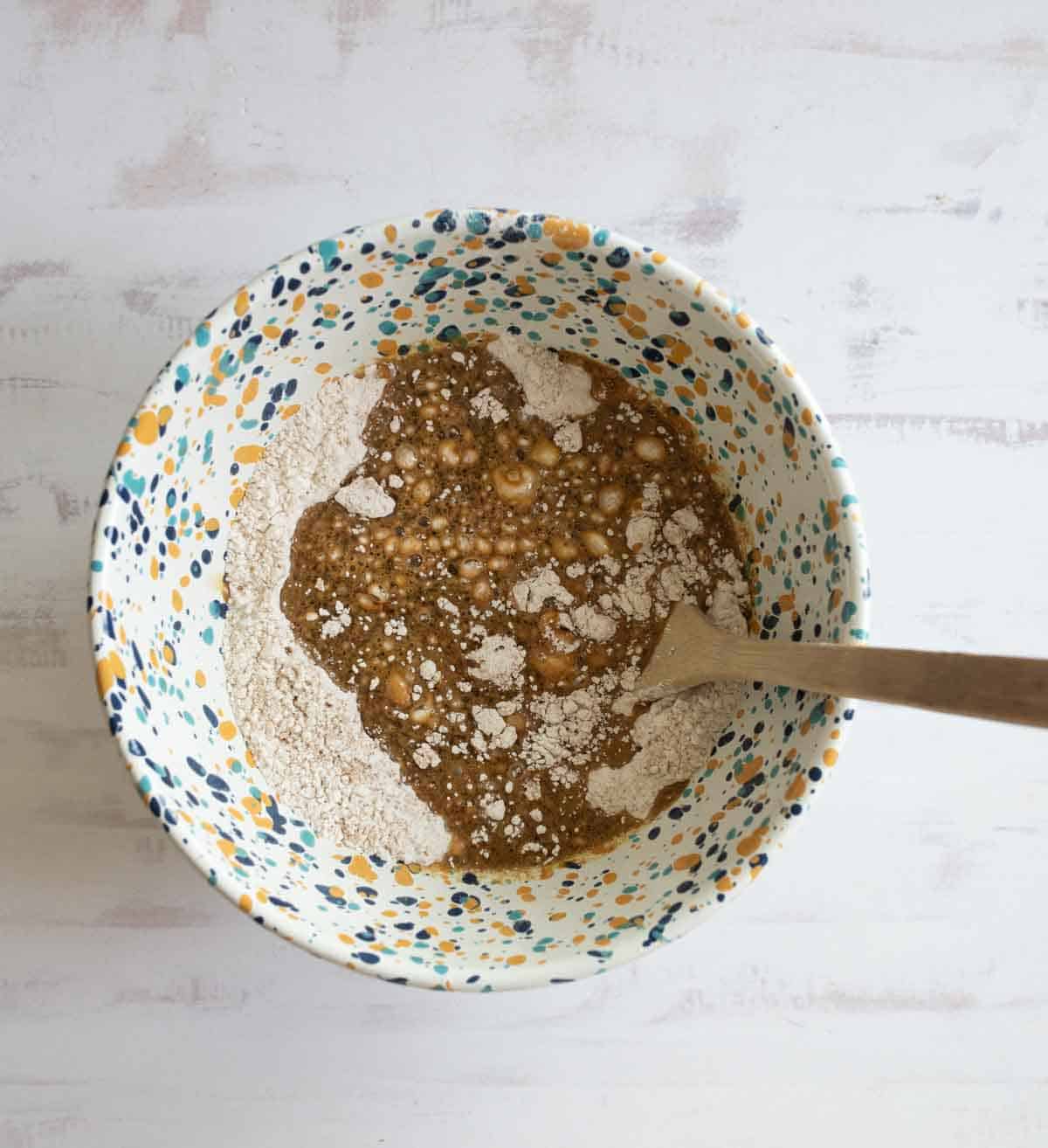 A colorful speckled bowl contains flour and brown liquid mixture with bubbles on the surface. A wooden spoon is partially submerged in the mixture, indicating the process of mixing ingredients. The background is a light textured surface.