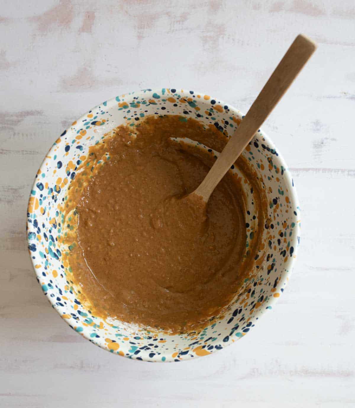 A speckled mixing bowl filled with creamy brown batter sits on a light wooden surface. A wooden spoon is partially submerged in the batter, leaning against the side of the bowl.