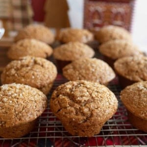 A batch of freshly baked gingerbread muffins topped with sugar crystals resting on a cooling rack. In the background, there are decorative, holiday-themed wooden trees and a red, house-shaped ornament.