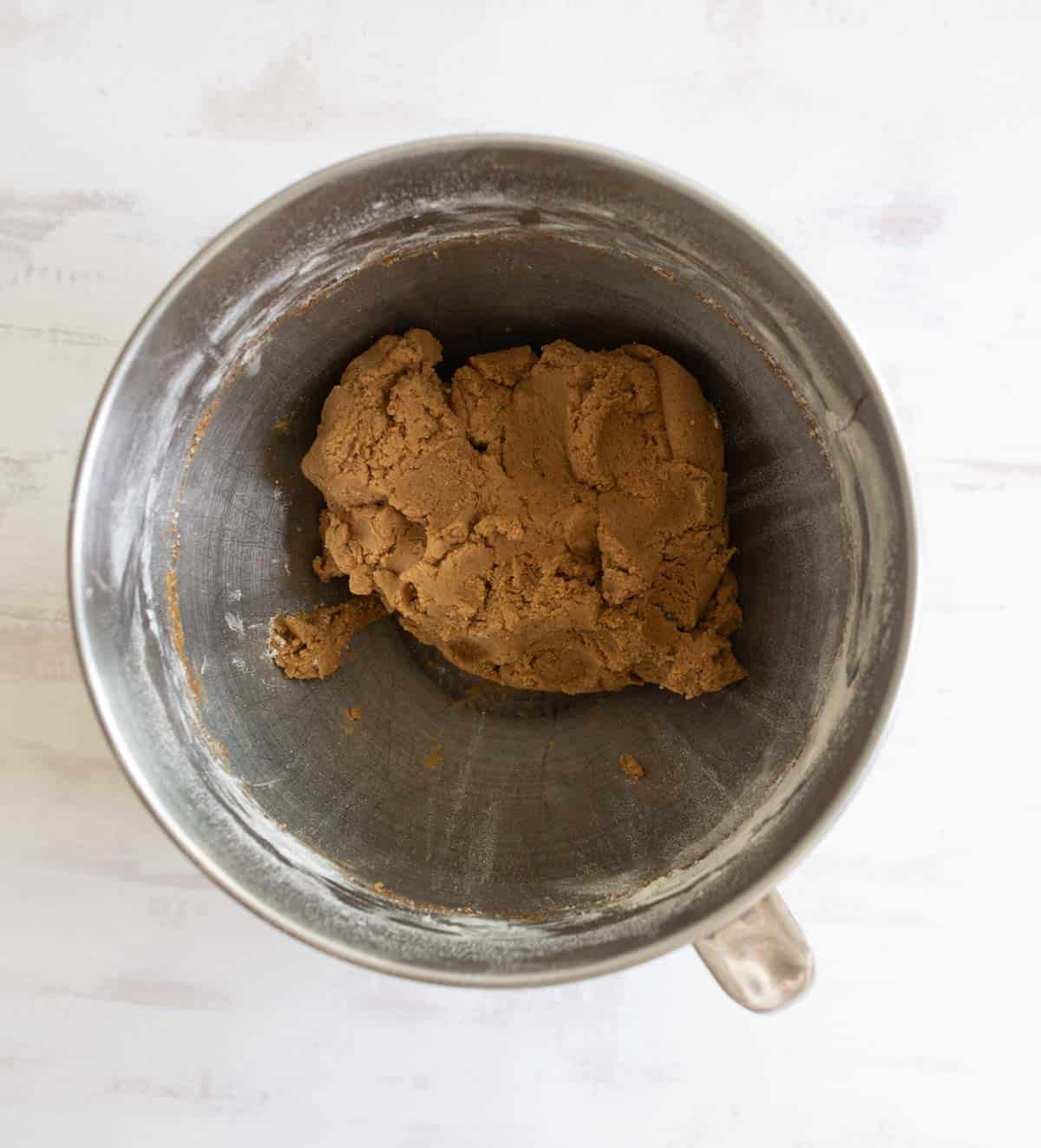 A metal mixing bowl containing a well-kneaded ball of brown dough sits on a light-colored surface. The dough appears smooth and evenly blended, likely for baking purposes.