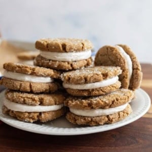 A plate of ginger molasses sandwich cookies with a creamy filling, stacked on a wooden table. The cookies are sprinkled with sugar, and a baking tray is visible in the background.