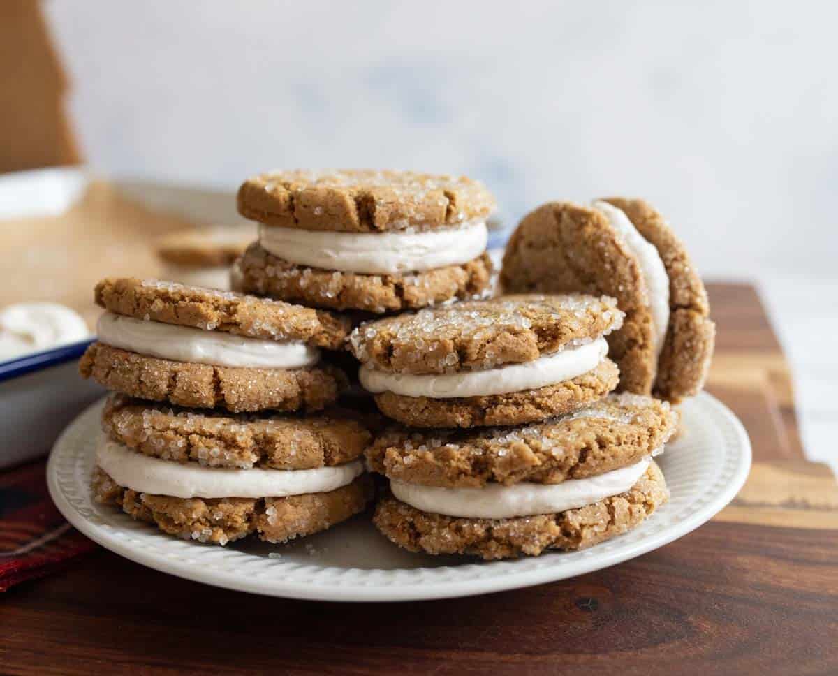 A plate of ginger molasses sandwich cookies with a creamy filling, stacked on a wooden table. The cookies are sprinkled with sugar, and a baking tray is visible in the background.