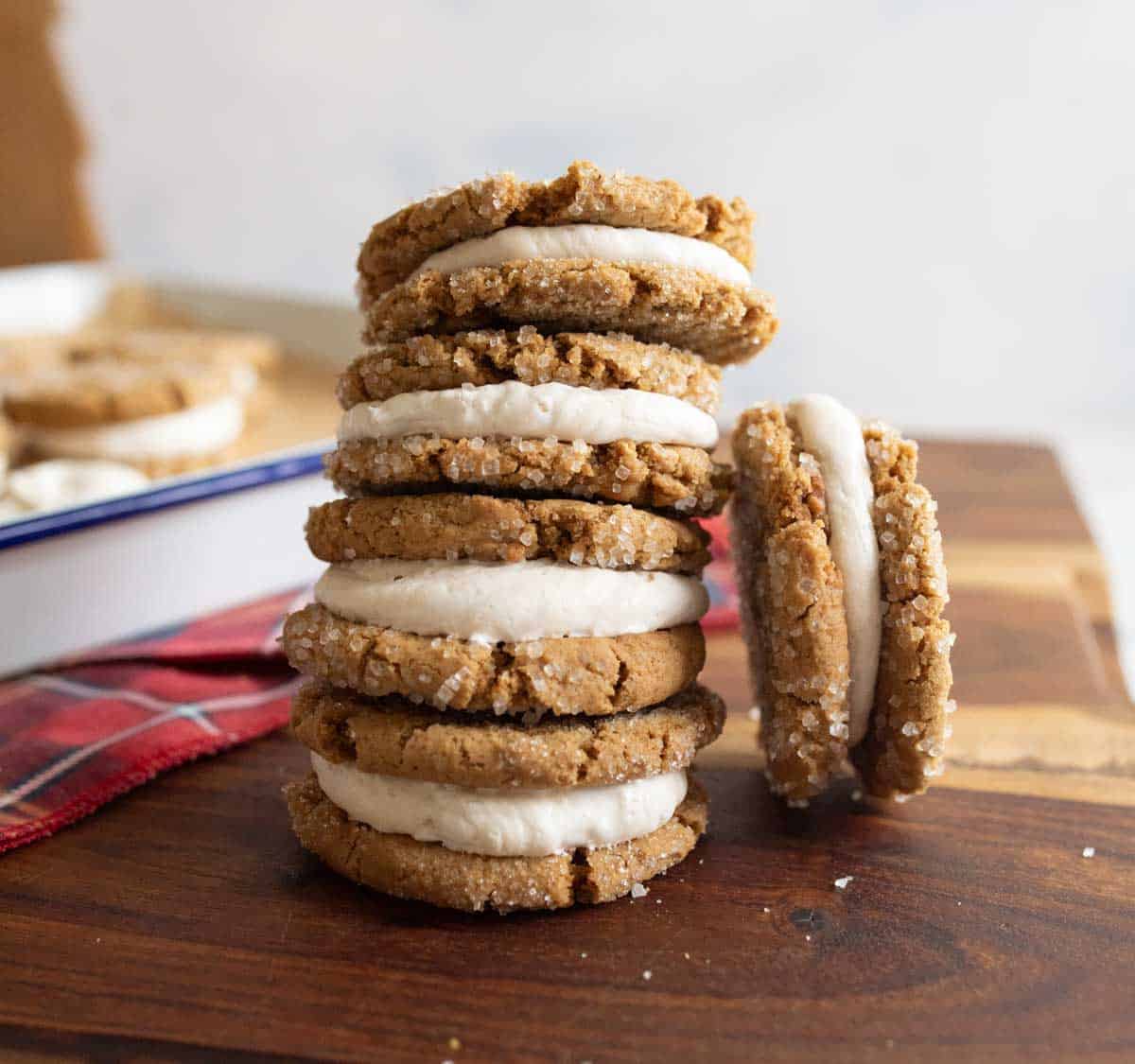 A stack of gingerbread sandwich cookies with white cream filling is arranged on a wooden surface. One cookie is leaning against the stack. A red plaid cloth is partially visible in the background.