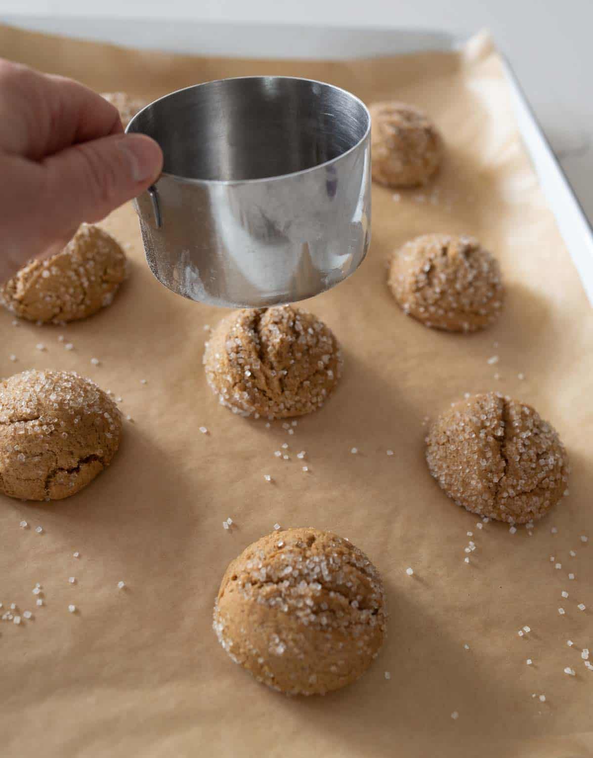 A hand holding a metal measuring cup gently presses down on freshly baked, sugar-coated cookies on a parchment-lined baking sheet. Several cookies remain unpressed, showcasing their cracked tops and coarse sugar texture.