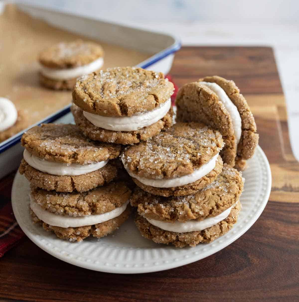 A plate of homemade sandwich cookies with creamy filling, sprinkled with sugar, displayed on a wooden table. In the background, more cookies rest on a baking tray.