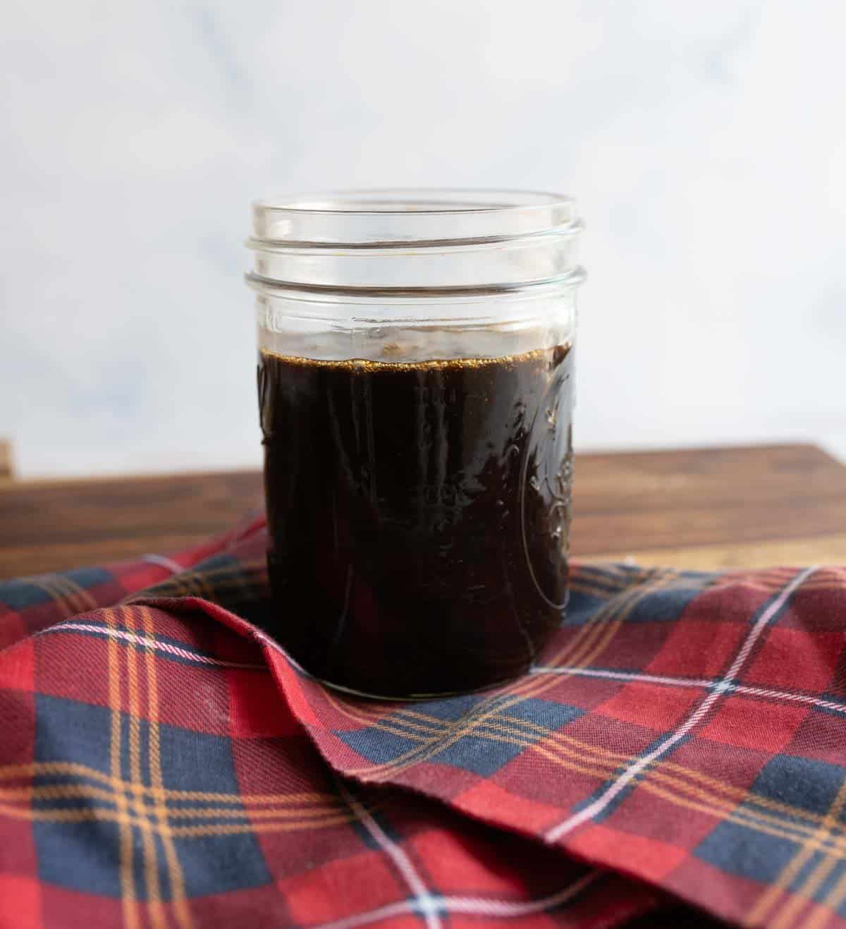 A glass jar filled with dark liquid sits on a folded red and blue plaid cloth. The jar is placed on a wooden surface, and the background is a soft white.