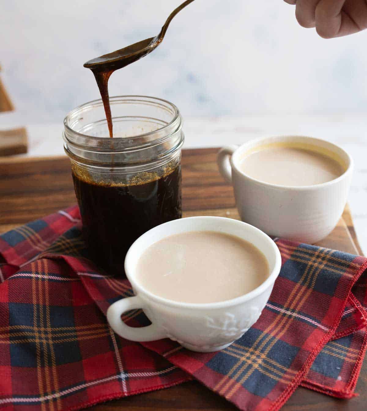 A jar of dark gingerbread syrup with a spoon above it, next to two cups of coffee with cream. The setup is on a wooden surface with a red plaid cloth underneath.