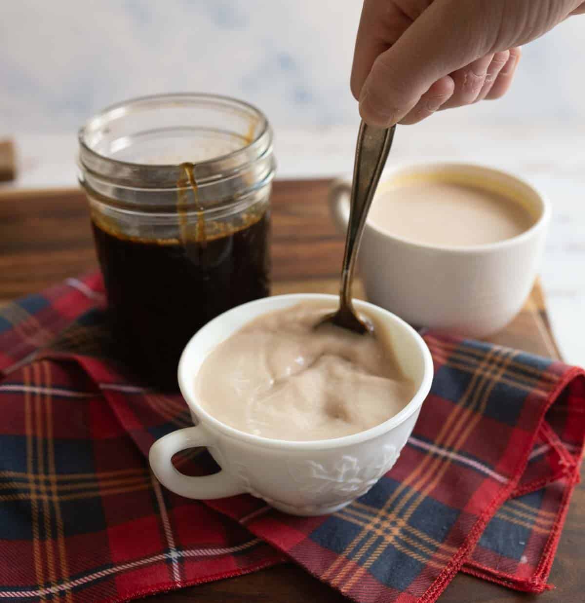 A hand stirs a creamy beverage in a white cup. Nearby, theres a jar of dark liquid and another white cup on a red and blue plaid cloth on a wooden surface.