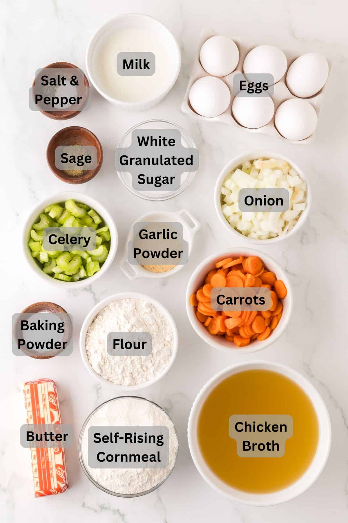 A delightful setup of cooking essentials is laid out on the marble surface, each labeled: eggs, milk, salt & pepper, sage, celery, white granulated sugar, onion, garlic powder, carrots, baking powder, flour, butter—all ready to craft a delicious cornbread dressing with self-rising cornmeal and chicken broth.