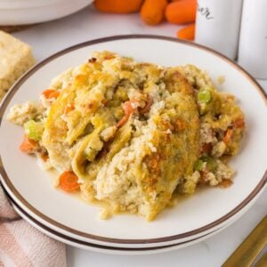 A plate of creamy chicken and stuffing casserole, featuring chunks of chicken, sliced carrots, and celery, topped with a golden, crispy crust. The dish is presented on a white plate with a dark rim. Other food ingredients are blurred in the background.