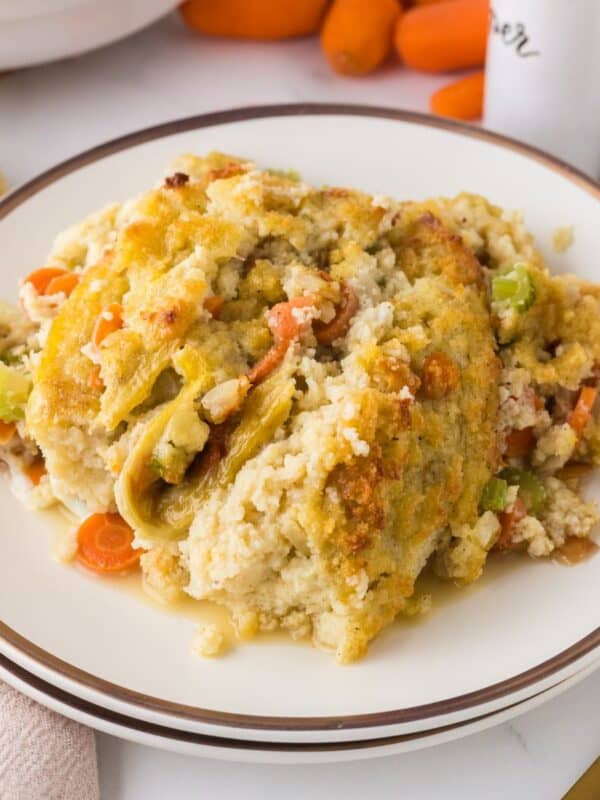 A plate of creamy chicken and stuffing casserole, featuring chunks of chicken, sliced carrots, and celery, topped with a golden, crispy crust. The dish is presented on a white plate with a dark rim. Other food ingredients are blurred in the background.