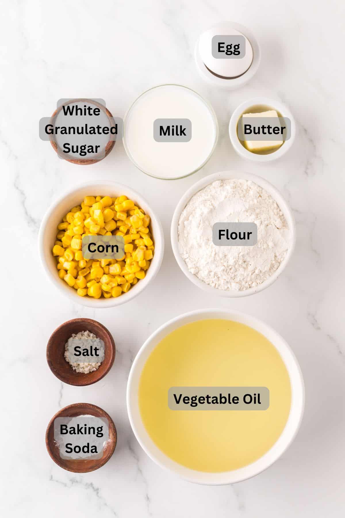 Top-down view of ingredients on a white marble surface, labeled: egg, milk, butter, flour, white granulated sugar, corn, salt, baking soda, and vegetable oil.